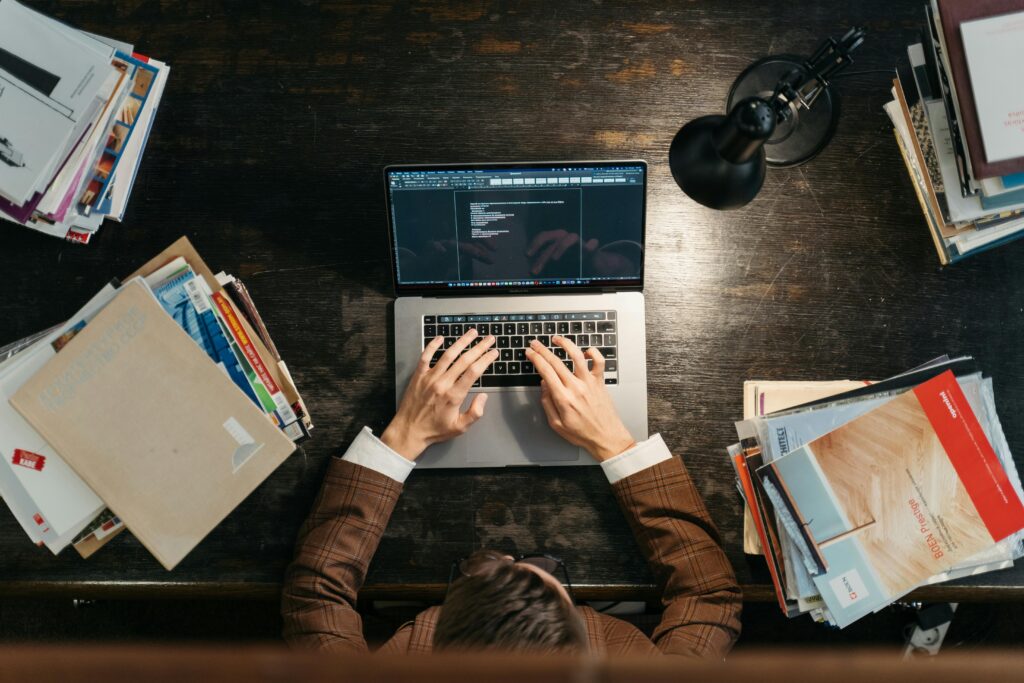 A person typing on a laptop at a cluttered desk with books and documents around.